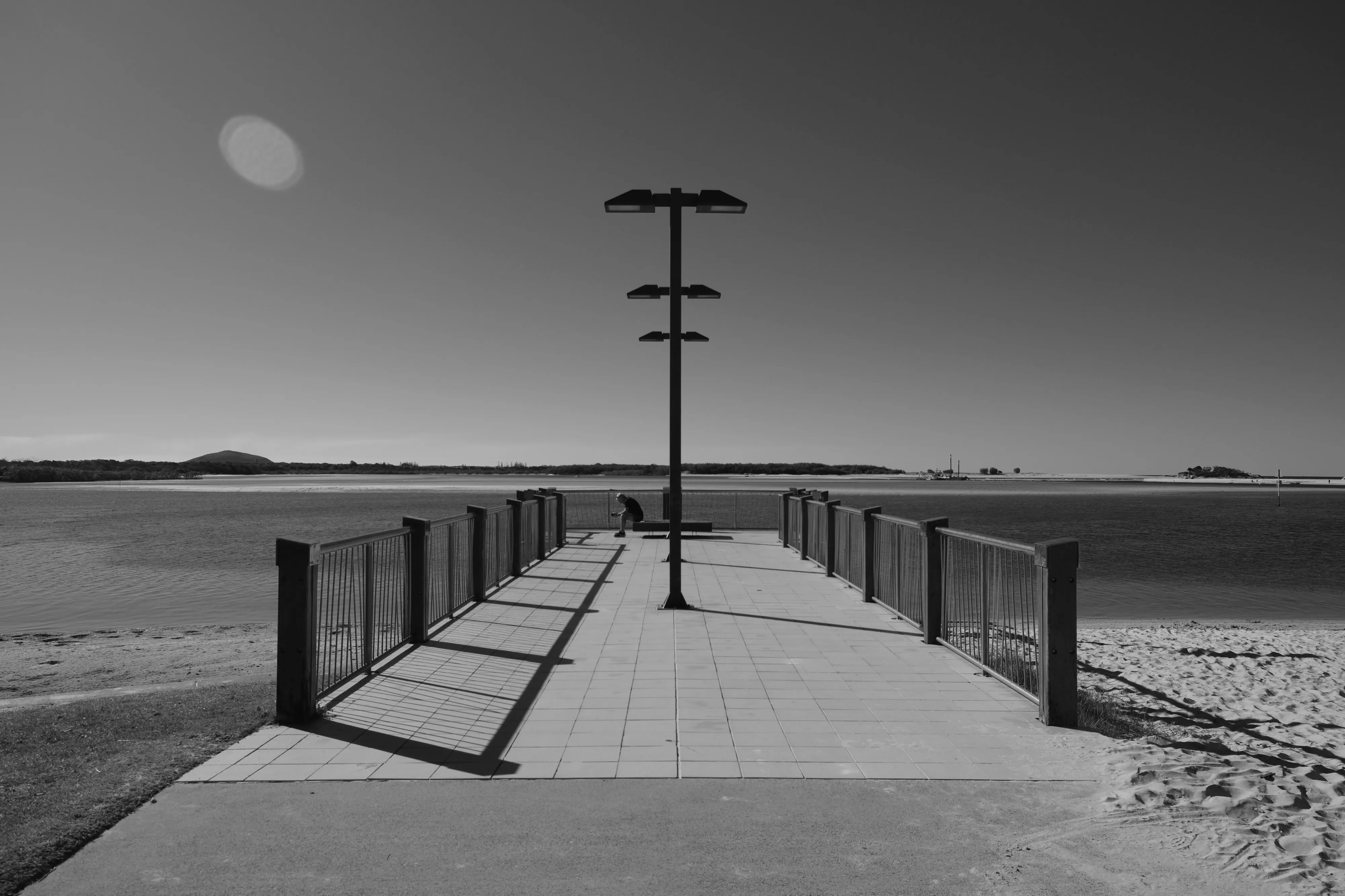 Lone person sitting on a bench at the end of a pier with a tall lamp post, shown in black and white against a wide sky and calm water