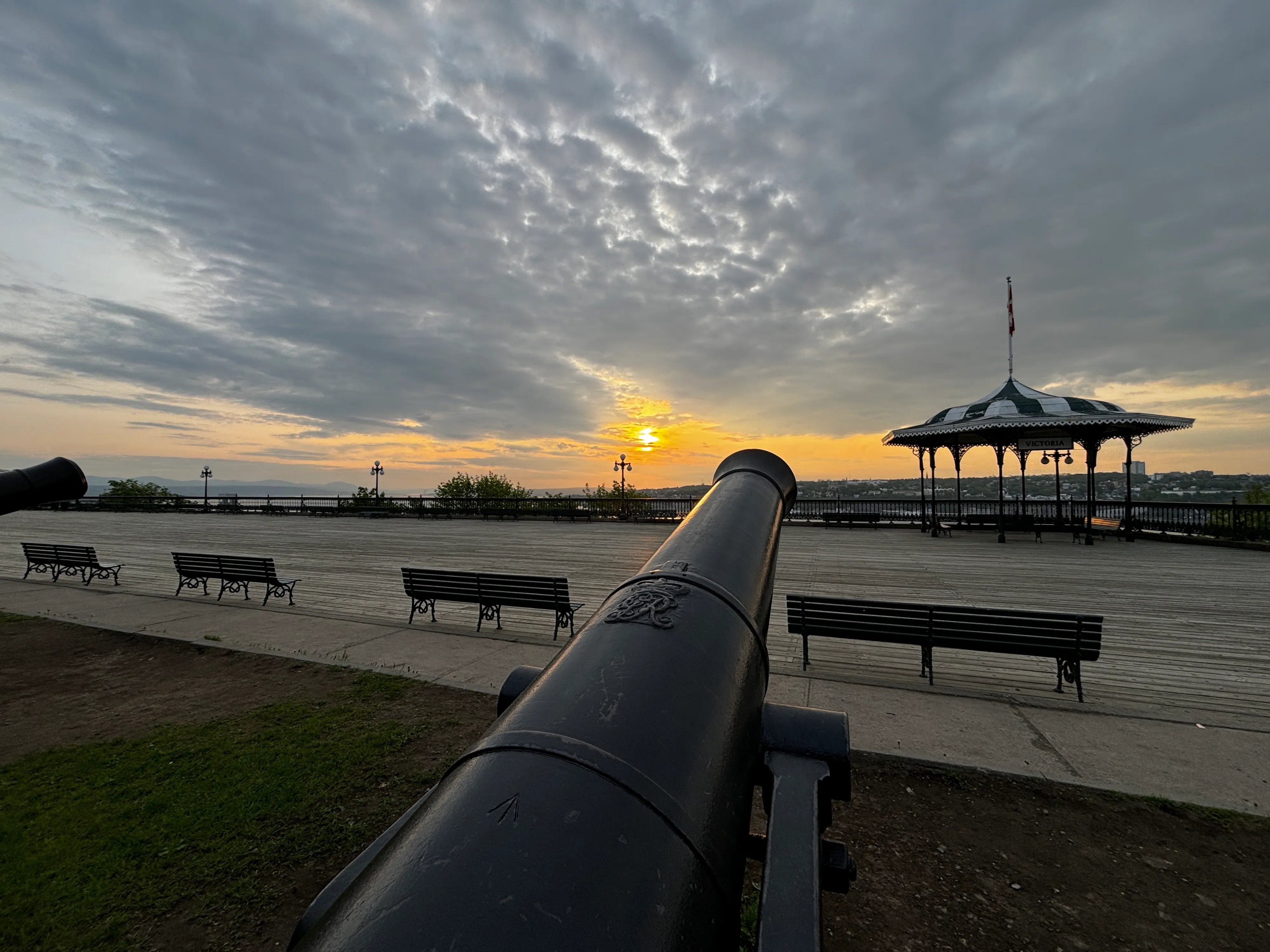 Old cannon pointing across a wooden promenade with benches and a bandstand, under a dramatic cloudy sky as the sun sets
