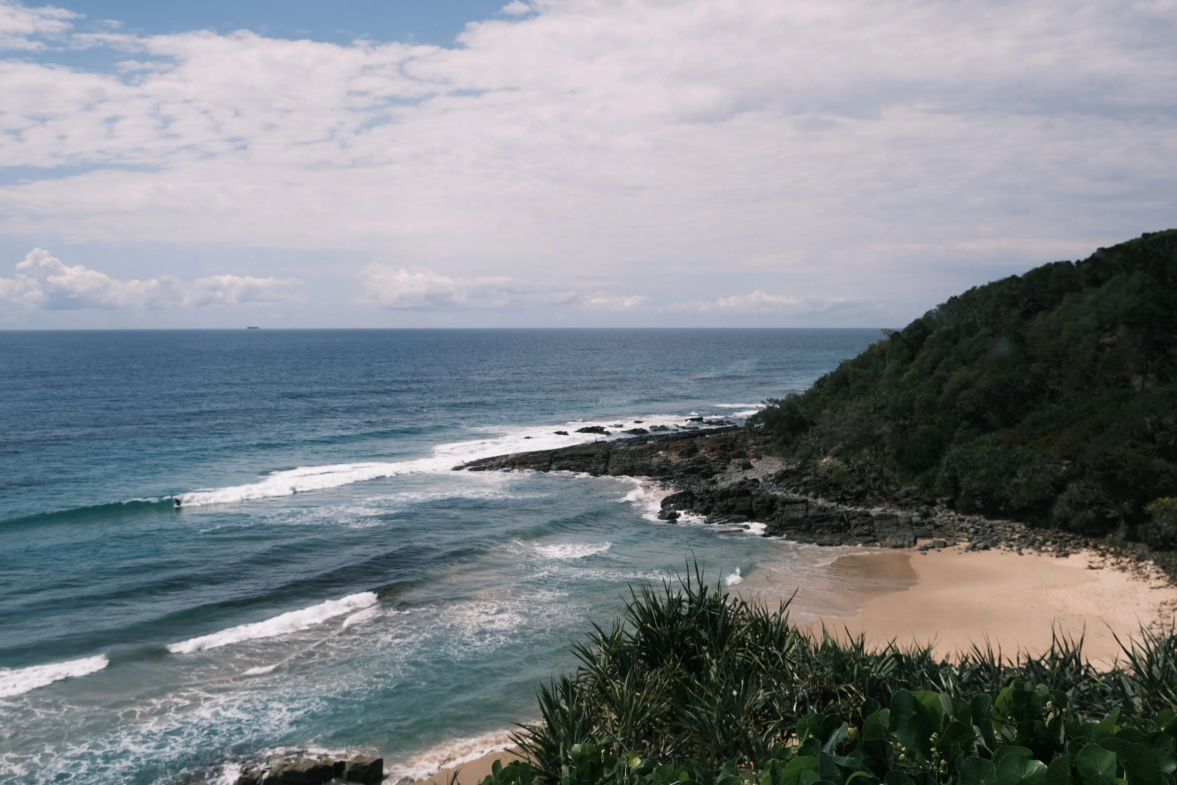 View from a headland over waves rolling onto a small beach, with a forested point and a cloudy sky
